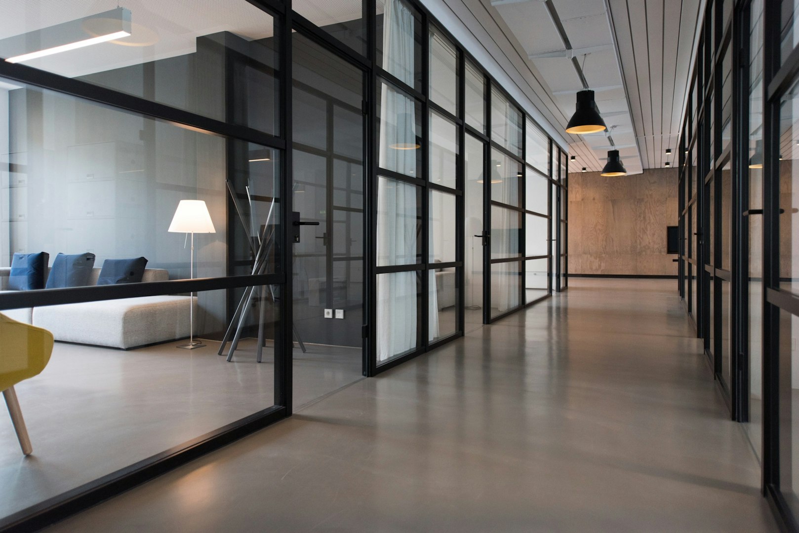 Modern office interior featuring large windows, strategic greenery, open workspace, and a seating area with a couch, stylish furniture, and a minimalist design.